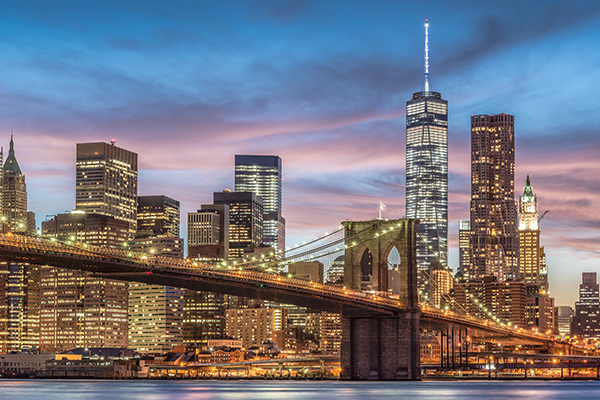 New York City skyline with the Brooklyn Bridge illuminated, a sunset in the background.